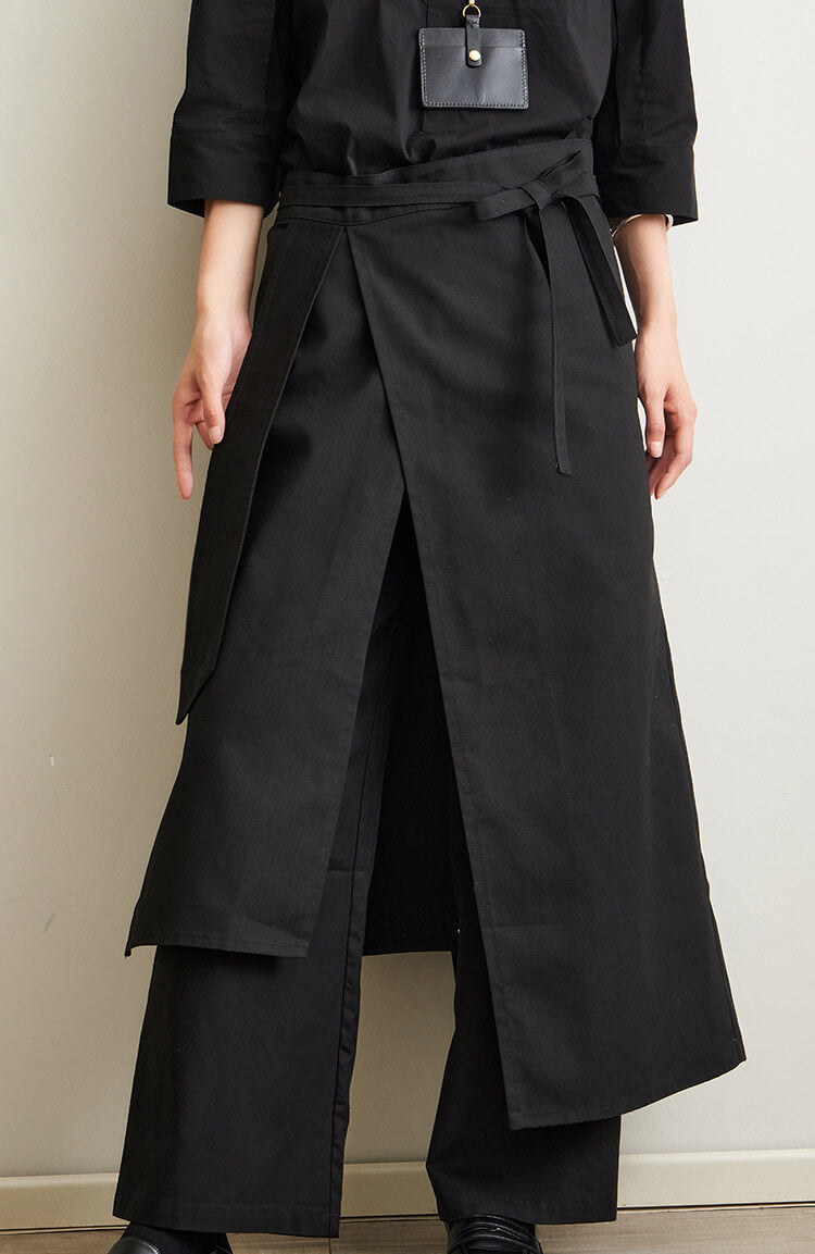 Person wearing a long black apron coat with wide-leg pants against a plain background