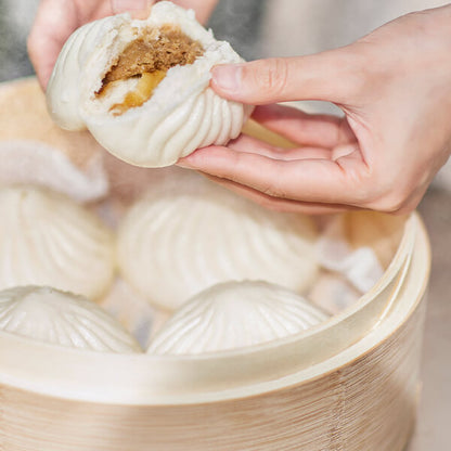 Person holding a steamed bun with a filling, with more buns in the background.
