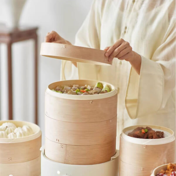 Person opening a bamboo steamer with food inside, set against a neutral background.